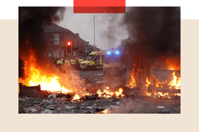 67a0ecc0-530b-11ef-8f0f-0577398c3339-png Colour image of riot police holding back protesters. In the foreground a police vehicle is engulfed by fire.