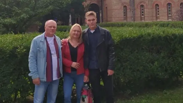 8c0feac0-def2-11ef-979f-05eee0cfa3a2-jpg Illia with his mum and dad. He is wearing a black leather jacket, his mum has shoulder-length blonde hair and is wearing blue jeans and a red jacket and his dad has short fair hair and is wearing a denim short and jeans. They are standing in front of a green bush with an old building behind them.