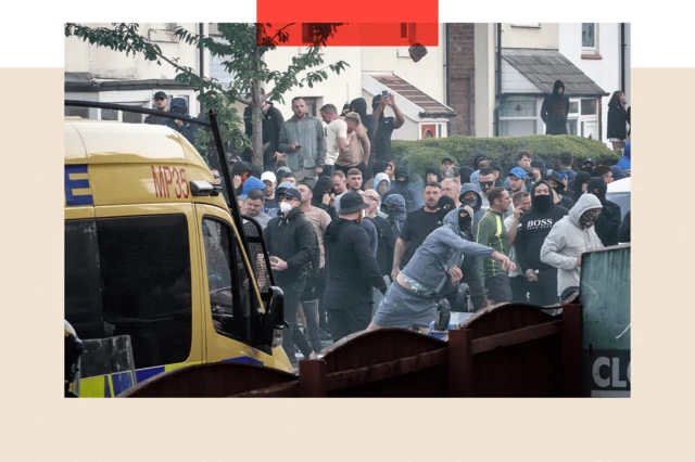 a1af9880-530b-11ef-8f0f-0577398c3339-png A crowd of male protesters behind a police van. One man in the foreground is wearing a black face covering and blue tracksuit top and shorts. He appears to be hurling something.