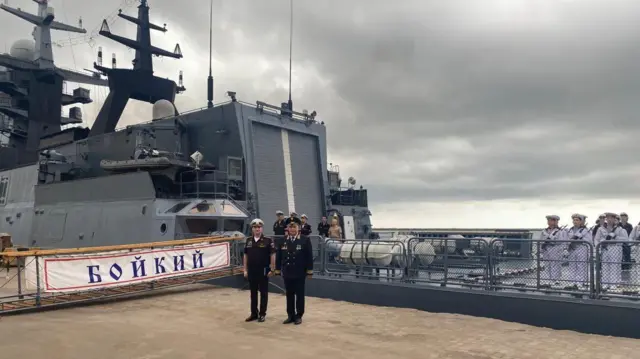 e87ed4e0-5113-11f0-918d-fb788d4dd5b5-jpg Russian naval officer standing on the deck of the Boikiy. A banner with the ships name can be seen in the background. Two officers are seen wearing uniforms, while enlisted men carry rifles.